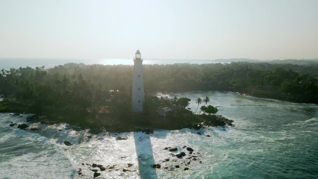 Drone Dondra lighthouse, southern tip of Sri Lanka, surrounded by greenery. Waves crash against rocks at tropical shoreline. Historic maritime landmark guides ships at Indian Ocean coast.
