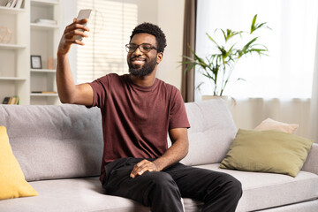 Happy African American Man Taking Selfie At Home. Cheerful Young Male With Glasses Enjoying Leisure Time On Couch With Smartphone. Indoor Lifestyle And Modern Technology Concept.