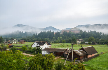 A tranquil morning in a rural village, misty mountains embrace traditional wooden houses amidst vibrant green fields. Verkhovyna, Carpathians, Ukraine
