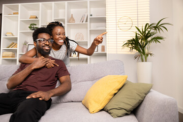 Happy Couple Relaxing On Sofa At Home, Pointing And Smiling, Cozy Modern Interior, Togetherness Concept