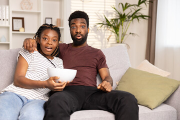 Surprised Afro Couple Watching TV On Couch With Snacks. Home Entertainment, Shock, Excitement, Leisure Time Concepts. Captured In Cozy Living Room. Perfect For Ads And Social Media Content.