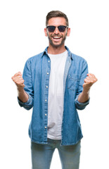 Young handsome man wearing sunglasses over isolated background celebrating surprised and amazed for success with arms raised and open eyes. Winner concept.