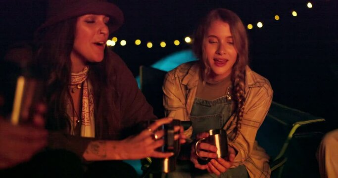 Night, camping and friends with coffee, laughing and talking on late outdoor adventure at tent. Campfire, hot drinks and women in dark at campsite for holiday, relax and happy chat in nature with cup