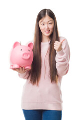 Young Chinese woman over isolated background holding piggy bank happy with big smile doing ok sign, thumb up with fingers, excellent sign