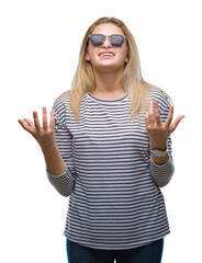 Young caucasian woman wearing sunglasses over isolated background crazy and mad shouting and yelling with aggressive expression and arms raised. Frustration concept.