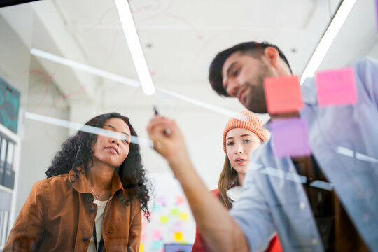 Business professional uses a glass wall covered with sticky notes to outline processes during a team meeting.