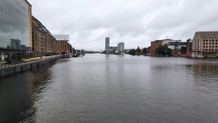 Cloudy cityscape with riverfront buildings and calm water under an overcast sky.