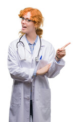 Young redhead woman wearing doctor uniform with a big smile on face, pointing with hand and finger to the side looking at the camera.
