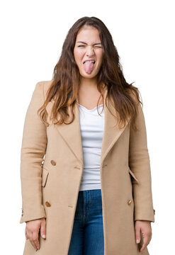 Beautiful plus size young woman wearing winter coat over isolated background sticking tongue out happy with funny expression. Emotion concept.