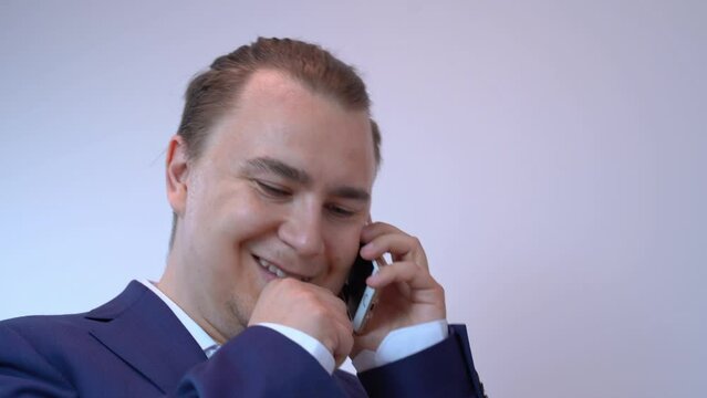 Portrait of male businessman in business suit, with stylish hairstyle, talking on phone, nodding head, smiling, bringing fist to mouth, coughing Positive communication, negotiations, successful deal