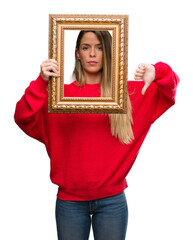Beautiful young woman holding vintage frame with angry face, negative sign showing dislike with...