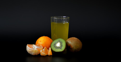 A glass of a mixture of tropical fruits with seeds on a black background, next to a ripe tangerine and kiwi.