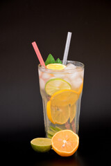 A tall faceted glass of refreshing lemonade with ice on a black background, next to pieces of ripe orange and lime.