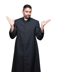 Young Christian priest over isolated background clueless and confused expression with arms and...