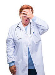 Senior caucasian doctor woman wearing medical uniform over isolated background peeking in shock covering face and eyes with hand, looking through fingers with embarrassed expression.