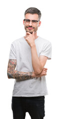 Young tattooed adult man looking confident at the camera with smile with crossed arms and hand raised on chin. Thinking positive.