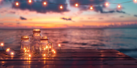 At dusk, a wooden table with mason jars and string lights overlooks the ocean. - obrazy, fototapety, plakaty