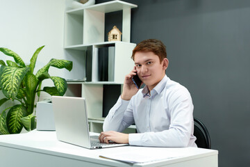 An ambitious man advises a client over the phone while doing work on a laptop in a modernly...