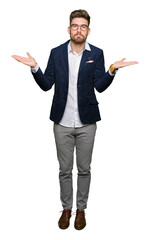 Young handsome business man wearing glasses clueless and confused expression with arms and hands raised. Doubt concept.
