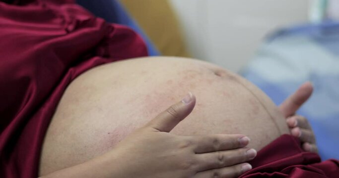 Pregnant woman wearing a pair of red silk pajamas is lying down on the bed as she is gently stroking her swollen belly with both of her hands.