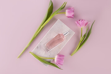 A chic bottle of women's perfume lies on a marble white podium among pink delicate tulips. Top view. Pink background. Beauty and fragrance concept.