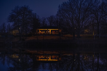 View of gazebo by lake in park in evening with silhouettes of people having party; everything reflects in calm water