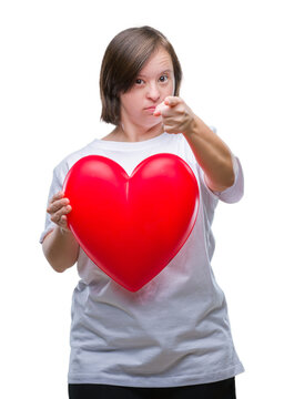 Young adult woman with down syndrome holding red heart over isolated background pointing with finger to the camera and to you, hand sign, positive and confident gesture from the front
