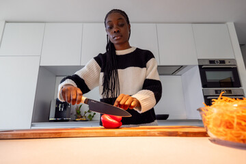 A young woman is pictured in a state of deep concentration while chopping a red bell pepper on a wooden cutting board. The kitchen's contemporary aesthetic is underscored by the simplicity and