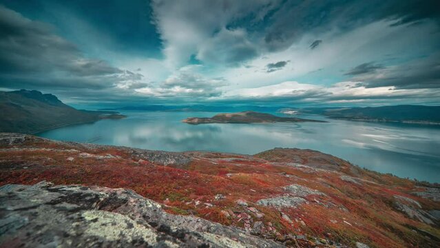 Whirling stormy clouds are backlit by the low sun over a calm mirror-like fjord and autumn tundra in the timelapse video.