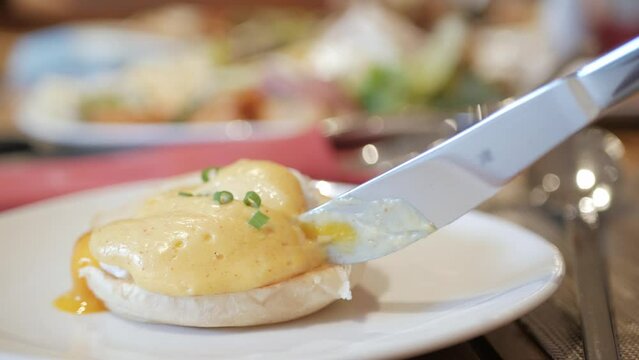 using knife and fork cutting egg yolk to eat egg benedict in white plate. Cutting poached egg with runny egg yolk over toast