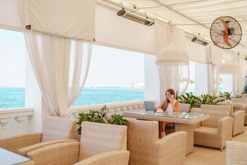 Woman coffee cafe laptop sea. Female freelancer using laptop on coffee break in modern cafe with sea view in the morning