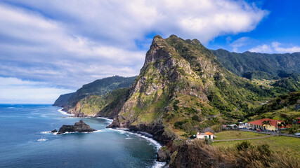 The cliff on Madeira rises above the azure ocean. The cliff is tall and steep, with an uneven and rocky surface. In the distance, waves crashing against the cliff's edge can be seen.