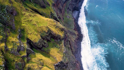 The coast of Madeira; green, rocky cliffs border with the white, frothy waves of the ocean, showcasing the contrast of natural elements.