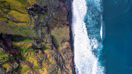 The coast of Madeira; green, rocky cliffs border with the white, frothy waves of the ocean, showcasing the contrast of natural elements.