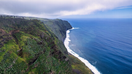The cliff on Madeira rises above the azure ocean. The cliff is tall and steep, with an uneven and rocky surface. In the distance, waves crashing against the cliff's edge can be seen.