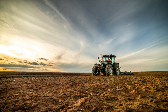 Wide-angle view of a tractor plowing farmland against a dramatic sunset sky