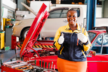 African woman mechanic standing, thumb up and hold a wrench with an opened radiator hood customer car in the background. Auto car repair service center. Professional service. Vehicle maintenance. - 781868780