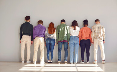 Diverse, silent group of seven people stand in row side by side facing white wall. Creative...