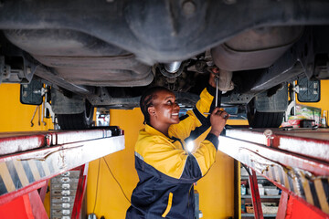 Portrait of an African female mechanic in yellow and blue uniform standing under the car bottom for inspecting in garage. Woman smiling while holding a wrench. Car repair service. Vehicle maintenance - 781868990