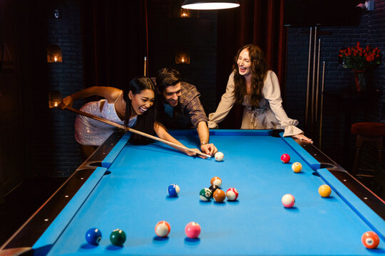 Young diversity group of people playing pool together with smile, enjoyment and fun. Young people spend time in billiards room at the nightclub. Men, women friends playing billiards. Nightlife concept