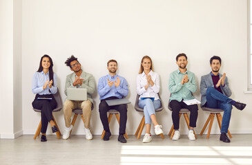 Fototapeta premium Audience clapping at a professional business conference. Group of several young diverse, multiracial people sitting in a row on chairs with laptops and clipboards on laps, clapping hands, and smiling