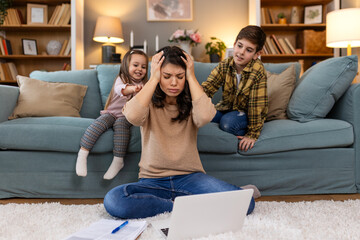Little naughty children distracting busy young mother from work on laptop at home, Son and daughter are making noises on sofa while mother is trying to work on her laptop