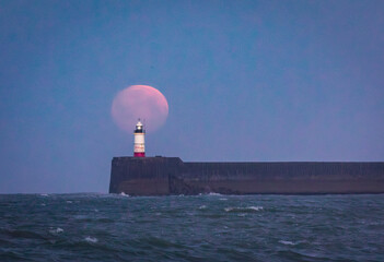 March full moon setting behind Newhaven lighthouse from Seaford beach on the east Sussex coast...