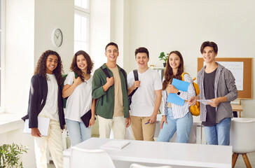 Portrait of a happy high school students group looking cheerful at camera and smiling standing in classroom. Guys and girls with backpacks on shoulders standing in a row. Back to school concept.
