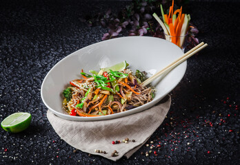 The image is of a bowl of stir-fried noodles, a popular Chinese dish. It includes various vegetables and is placed on a table indoors. Ingredients include noodles, vegetables, and meat or tofu.