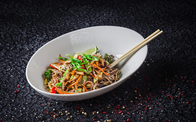 The image is of a bowl of stir-fried noodles, a popular Chinese dish. It includes various vegetables and is placed on a table indoors. Ingredients include noodles, vegetables, and meat or tofu.