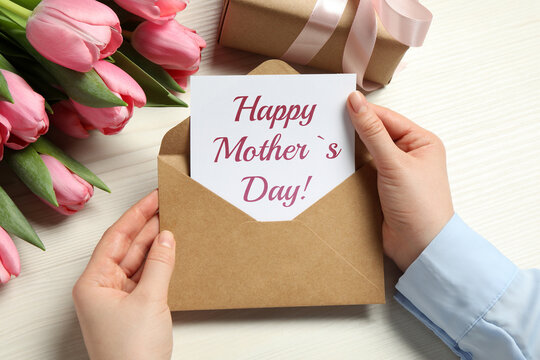 Happy Mother's Day. Woman taking greeting card out of envelope, closeup. Gift box and bouquet of beautiful flowers on white wooden table