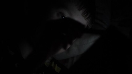Caucasian boy 9 years old uses a tablet computer in the dark using a stylus while lying in bed. A schoolboy uses a gadget before going to bed.