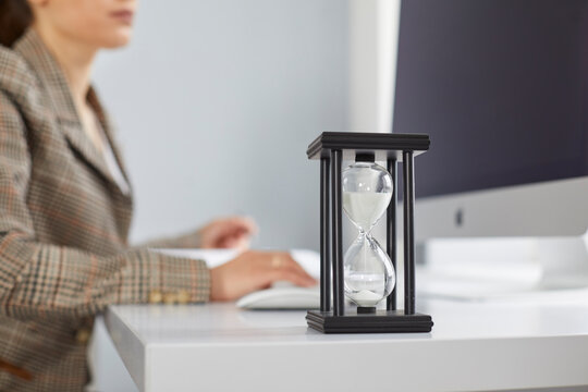 Close up of sand glass on desk of busy office worker trying to meet deadline. Closeup of hour glass with woman sitting at table and working on desktop computer in background. Time and business concept