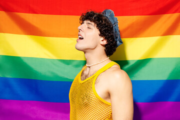 Side profile view young calm cool happy gay Latin man he wearing mesh tank top hat clothes close eyes stand on striped rainbow flag background studio portrait. Pride day June month love LGBT concept. - Powered by Adobe